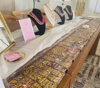 Bright indoor jewelry display on a wooden table in a home entryway: busts showcasing layered necklaces, racks of gold chains and clear organizers filled with charms, earrings and beads.