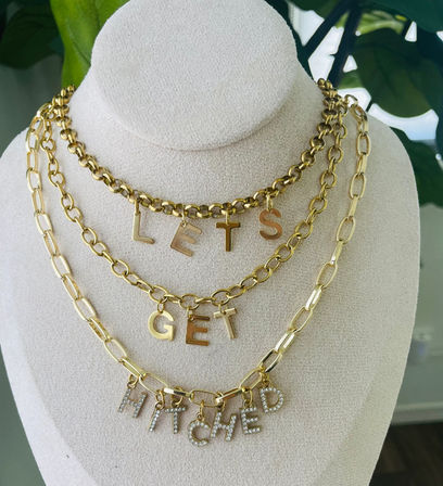Playful layered gold chain necklaces on a cream display bust spelling LETS GET HITCHED with sparkling rhinestone letter charms, styled as a bridal statement accessory