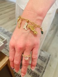 Close-up of a hand wearing a gold charm bracelet with glitter sunglasses, enamel cowboy boot, miniature motorcycle and cactus charms, paired with a gold statement ring and French-tip nails.