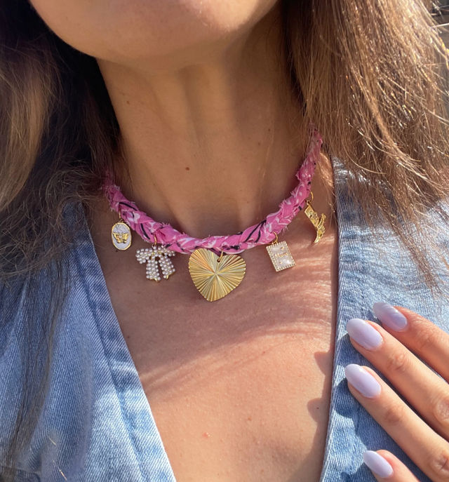 Sunlit close-up of a person's neckline wearing a pink braided choker with gold charms — textured heart pendant, pearl bow, square charm and small medallion — paired with a light denim top and pastel lavender nails.