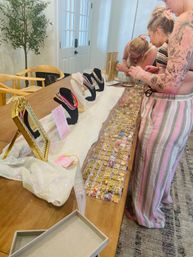 Home pop-up jewelry display on a wooden dining table with necklace busts, mirror and clear trays of gold charms and colorful beads as two people browse