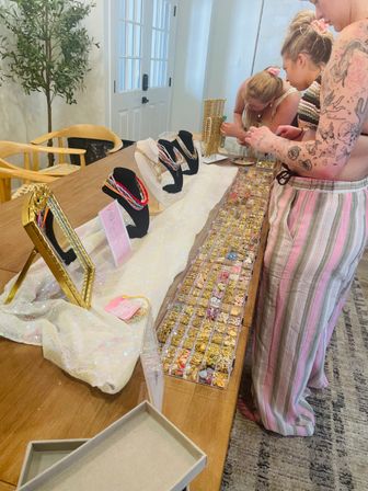 Home pop-up jewelry display on a wooden dining table with necklace busts, mirror and clear trays of gold charms and colorful beads as two people browse