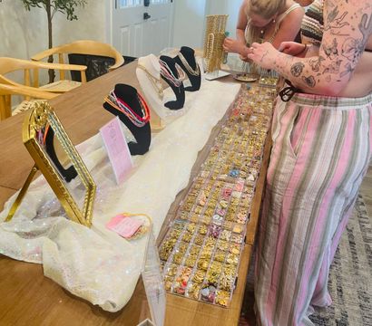 Home pop-up jewelry display on a wooden dining table with necklace busts, mirror and clear trays of gold charms and colorful beads as two people browse