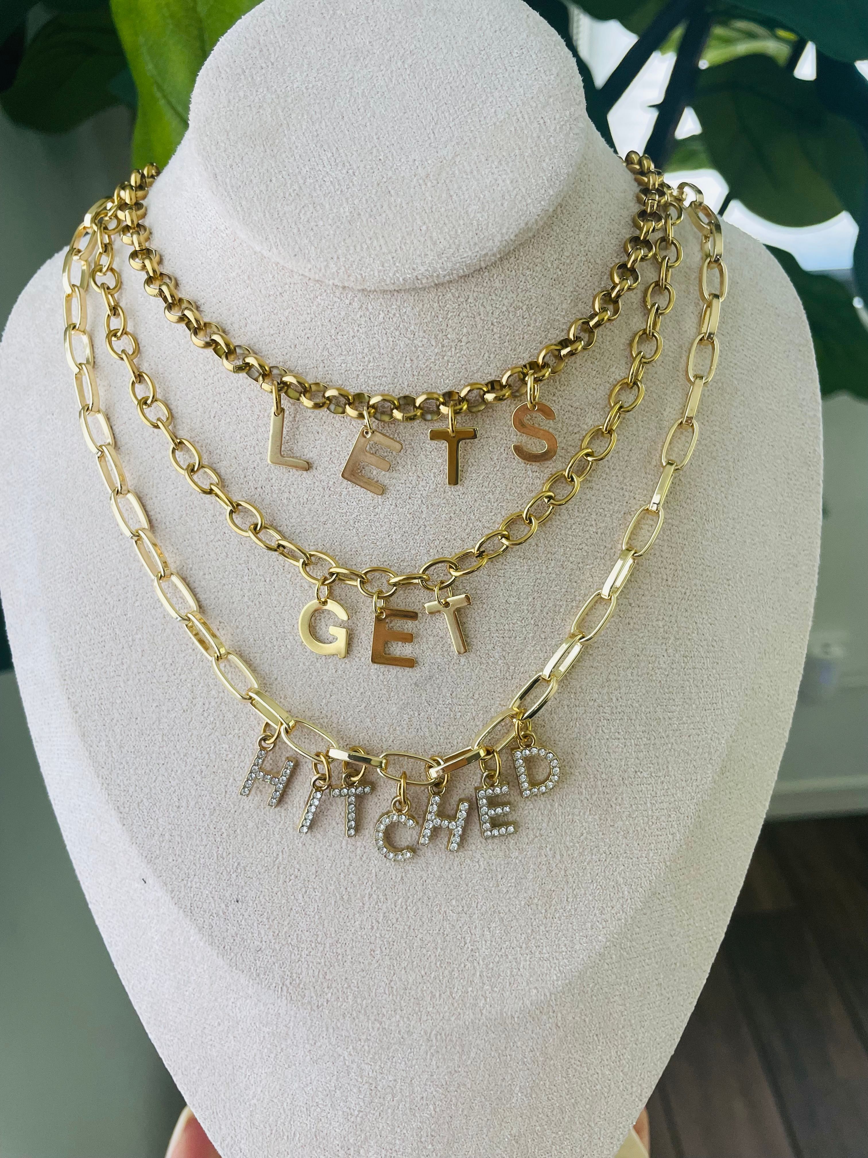 Playful layered gold chain necklaces on a beige display bust spelling LETS GET HITCHED with rhinestone-encrusted bottom letters — bridal statement charm necklace, wedding jewelry display