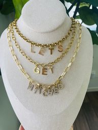 Playful layered gold chain necklaces on a beige display bust spelling LETS GET HITCHED with rhinestone-encrusted bottom letters — bridal statement charm necklace, wedding jewelry display