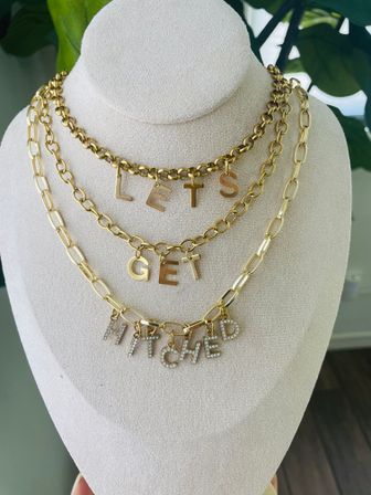 Playful layered gold chain necklaces on a beige display bust spelling LETS GET HITCHED with rhinestone-encrusted bottom letters — bridal statement charm necklace, wedding jewelry display