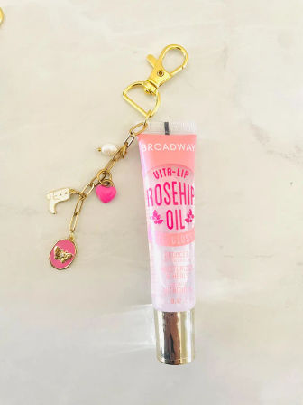 Pink rosehip oil lip gloss tube with metallic cap attached to a gold charm keychain featuring a pink heart, butterfly medallion and faux pearl on a light marble surface