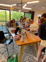 People gathered around a wooden table at a home craft party assembling jewelry — bead trays, necklace displays, star balloons and large windows with green trees outside.