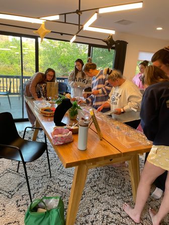 People gathered around a wooden table at a home craft party assembling jewelry — bead trays, necklace displays, star balloons and large windows with green trees outside.