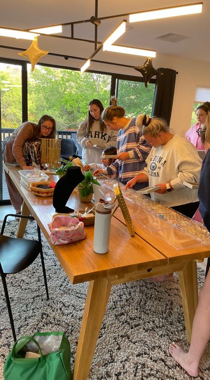 People gathered around a wooden table at a home craft party assembling jewelry — bead trays, necklace displays, star balloons and large windows with green trees outside.