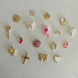 Flatlay of playful assorted gold and enamel jewelry charms — hearts, key, bow, cross, horseshoe, chili pepper, pearl bow and pink boot — on white marble surface