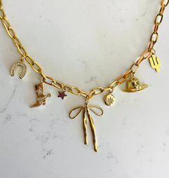 Gold charm necklace on white marble background — chunky chain with Southwestern-inspired charms: sparkly horseshoe, cowboy boot with spur, small star, bow pendant, engraved star medallion, cowboy hat and cactus.
