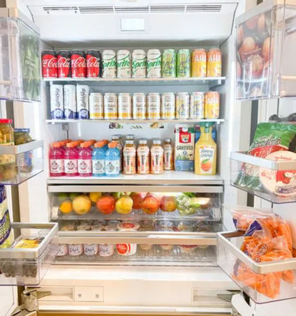Open kitchen refrigerator neatly stocked with brightly colored canned sodas and sparkling water, bottled juices and sports drinks, yogurt cups, fresh apples and oranges, and packaged snacks in the door.
