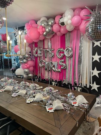 Indoor girls' party setup with pink and silver balloon garland, "LET'S GO GIRLS" foil letters, pink streamer backdrop and disco-ball centerpieces and party favors on a wooden table.