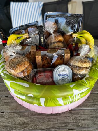 Poolside brunch gift basket on a wooden deck filled with bagels and seeded rolls, muffins, fresh bananas, mixed fruit, a tub of cream cheese and a mini sparkling wine bottle.