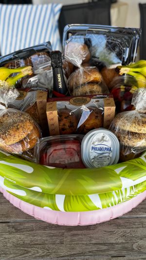 Poolside brunch gift basket on a wooden deck filled with bagels and seeded rolls, muffins, fresh bananas, mixed fruit, a tub of cream cheese and a mini sparkling wine bottle.