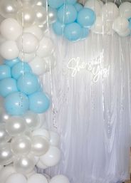 Indoor engagement party backdrop with pastel blue, white and pearlescent balloon arch, sheer white drape, silver curling ribbons and neon "She said Yes!" sign