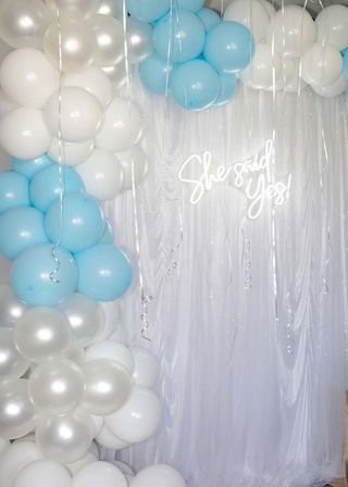 Indoor engagement party backdrop with pastel blue, white and pearlescent balloon arch, sheer white drape, silver curling ribbons and neon "She said Yes!" sign