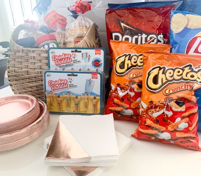 Playful party snack spread on a white kitchen counter: wicker basket with bread and peanut butter, large bags of chips (Cheetos, Doritos, Lay’s), rose-gold paper plates and napkins, and novelty "Drinking Buddies" drink markers.