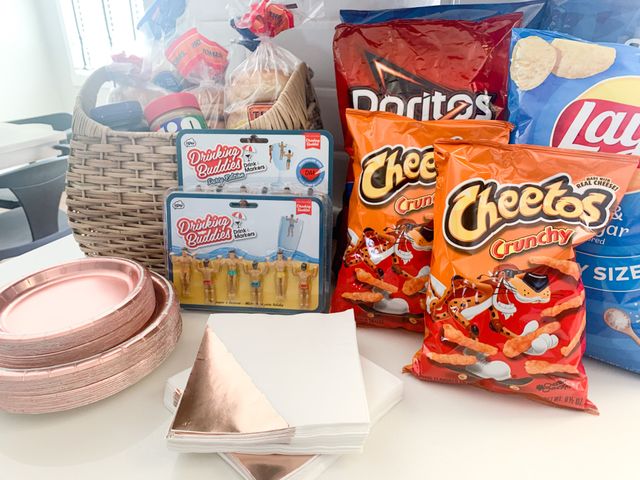 Playful party snack spread on a white kitchen counter: wicker basket with bread and peanut butter, large bags of chips (Cheetos, Doritos, Lay’s), rose-gold paper plates and napkins, and novelty "Drinking Buddies" drink markers.