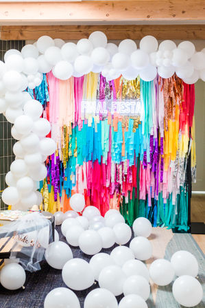 Indoor party photo backdrop with a white balloon arch and scattered white balloons framing a vibrant rainbow tassel wall and neon FINAL FIESTA sign