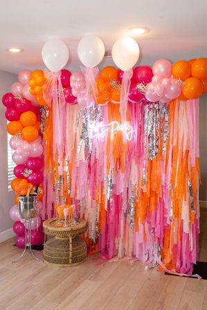 Indoor photo booth backdrop with pink, orange and white balloon garland, cascading crepe streamers and neon "Let's Party" sign, wicker table with champagne bucket and pink flutes.