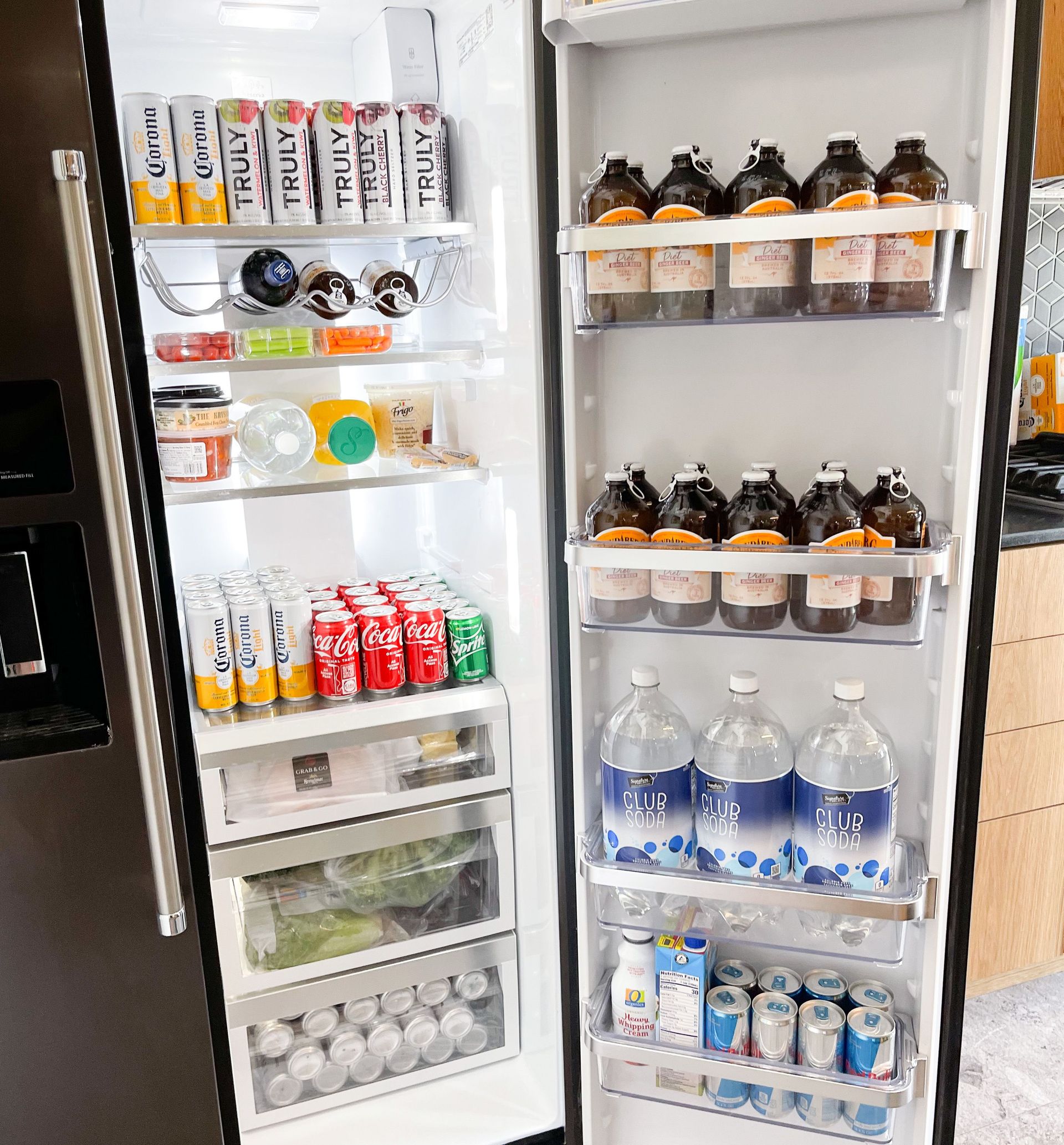 Open modern stainless-steel fridge neatly stocked with beverages and produce: tall seltzer/beer cans, rows of Coca‑Cola and Sprite cans, amber glass growlers on door shelves, three large club soda bottles and produce drawers.