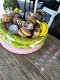 Inflatable brunch gift basket on a wooden patio table with bagels, cream cheese tub, muffins, bananas, mixed fruit and a bottle of sparkling wine, plus lined plastic champagne flutes