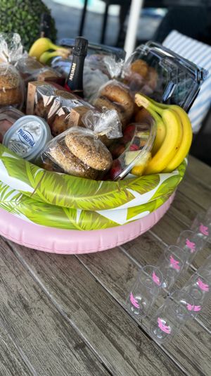 Inflatable brunch gift basket on a wooden patio table with bagels, cream cheese tub, muffins, bananas, mixed fruit and a bottle of sparkling wine, plus lined plastic champagne flutes