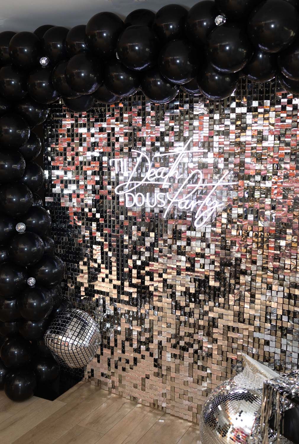 Glam party photo wall with mirrored sequin backdrop, oversized black balloon arch, shiny disco balls and white neon script sign — stylish photo booth setup on wooden floor