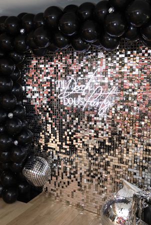 Glam party photo wall with mirrored sequin backdrop, oversized black balloon arch, shiny disco balls and white neon script sign — stylish photo booth setup on wooden floor