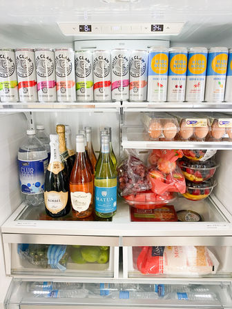 Organized kitchen refrigerator interior: top shelf lined with canned hard seltzers and vodka sodas, middle shelf holding champagne and wine bottles, egg cartons and stacked fruit bowls, lower drawers with limes and shredded cheese, and bottled water in the door.