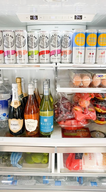 Organized kitchen refrigerator interior: top shelf lined with canned hard seltzers and vodka sodas, middle shelf holding champagne and wine bottles, egg cartons and stacked fruit bowls, lower drawers with limes and shredded cheese, and bottled water in the door.