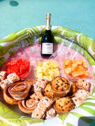 Poolside floating tray holding a chilled champagne bottle, clear plastic flutes, bowls of watermelon, pineapple and cantaloupe, and assorted pastries and muffins on a sunny summer day.