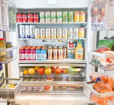 Open modern kitchen refrigerator neatly stocked with canned sodas and sparkling water on top shelves, bottled juices and milk alternative, fresh apples and citrus in the crisper, yogurts and snacks on door shelves.