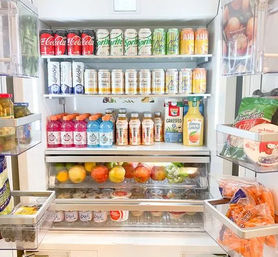 Open modern kitchen refrigerator neatly stocked with canned sodas and sparkling water on top shelves, bottled juices and milk alternative, fresh apples and citrus in the crisper, yogurts and snacks on door shelves.