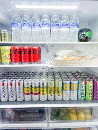Organized refrigerator shelf stocked with bottled water on top, red soda cans, rows of assorted hard seltzers and vodka seltzers, a covered sushi tray, and a drawer of lemons and limes.