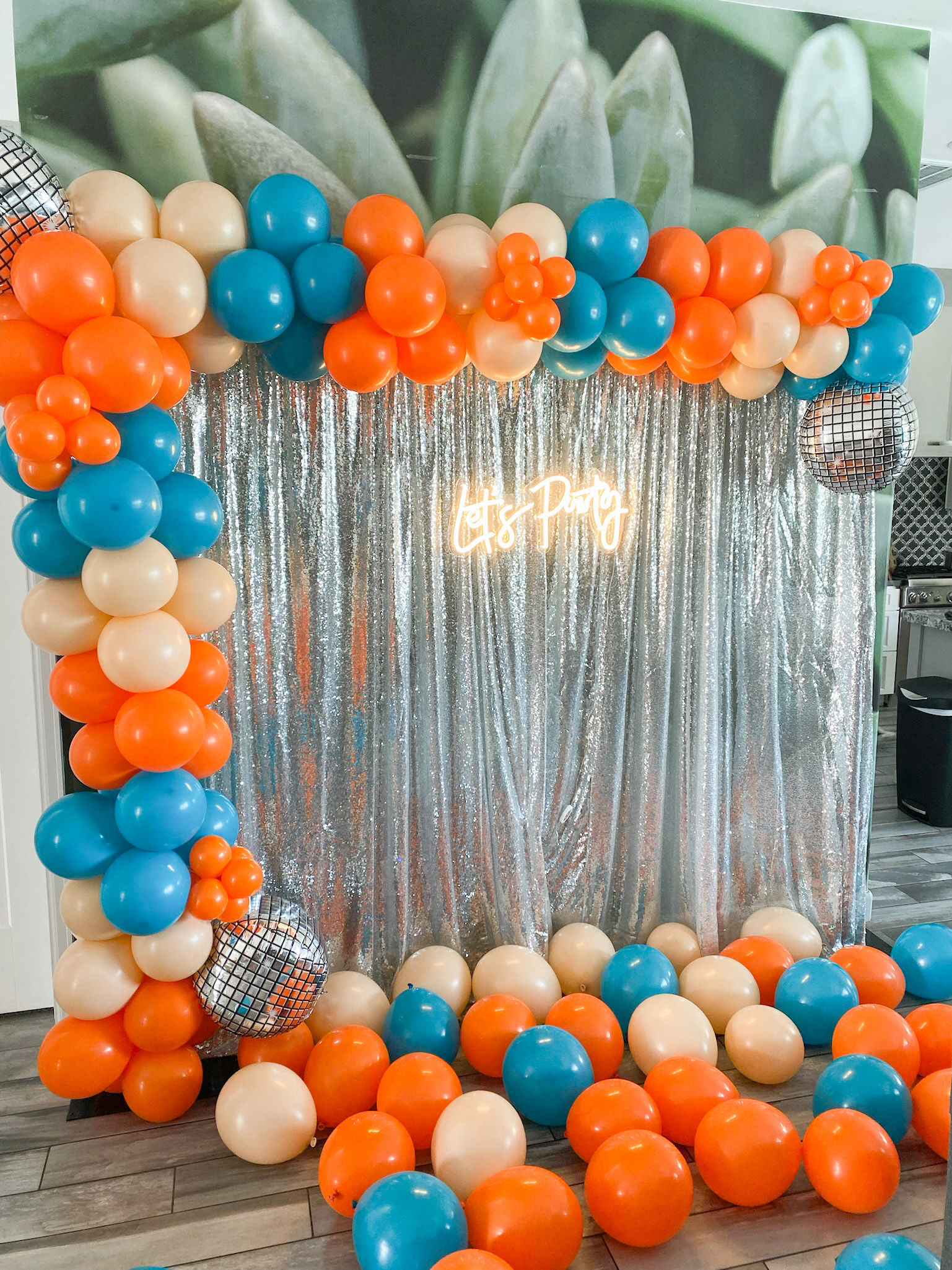 Indoor party photo backdrop with silver sequin curtain, neon “Let’s Party” sign, orange, teal and cream balloon arch and scattered balloons with mirrored disco balls