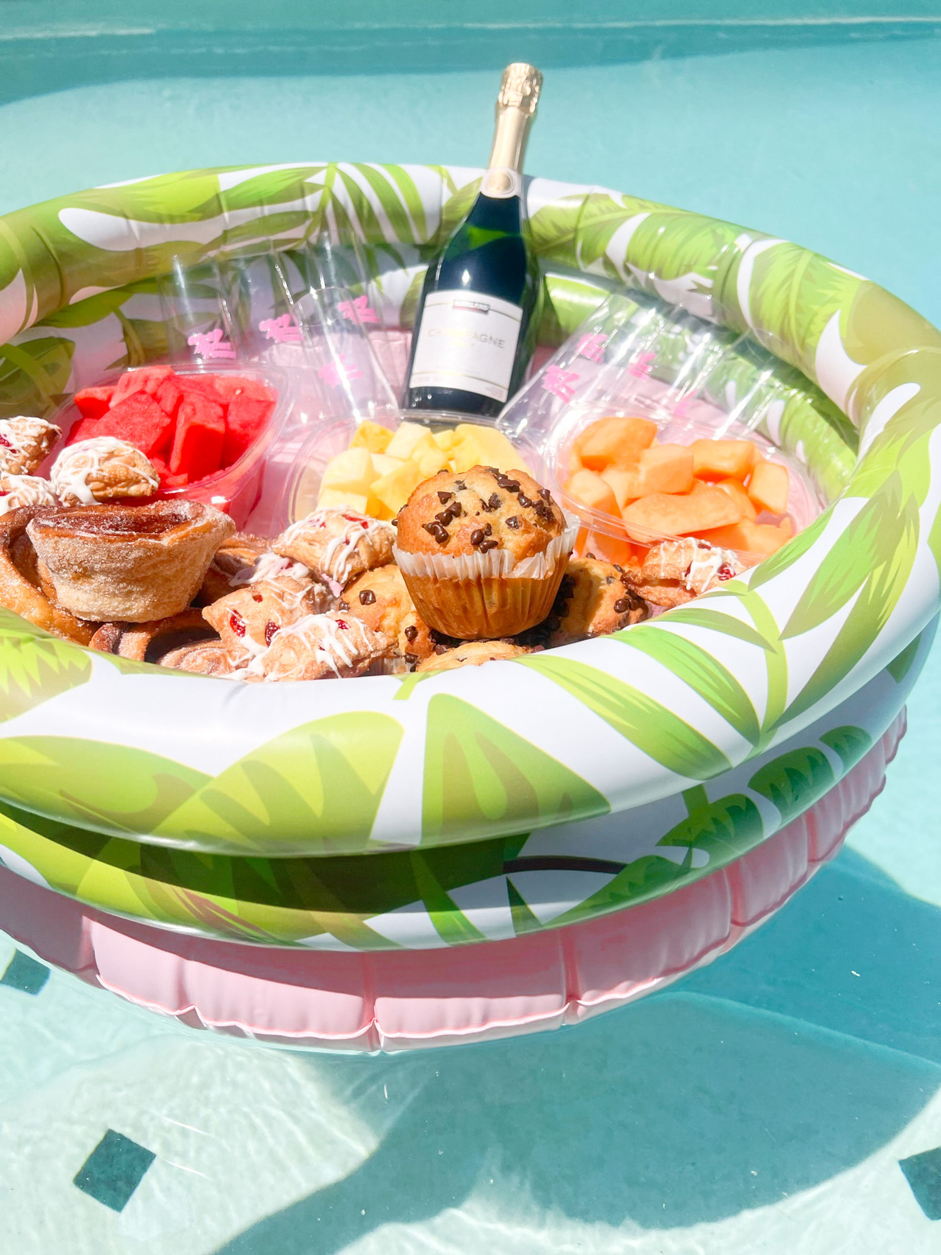 Tropical inflatable floating tray in a pool with champagne, muffins, pastries and bowls of watermelon, pineapple and cantaloupe — summer poolside brunch