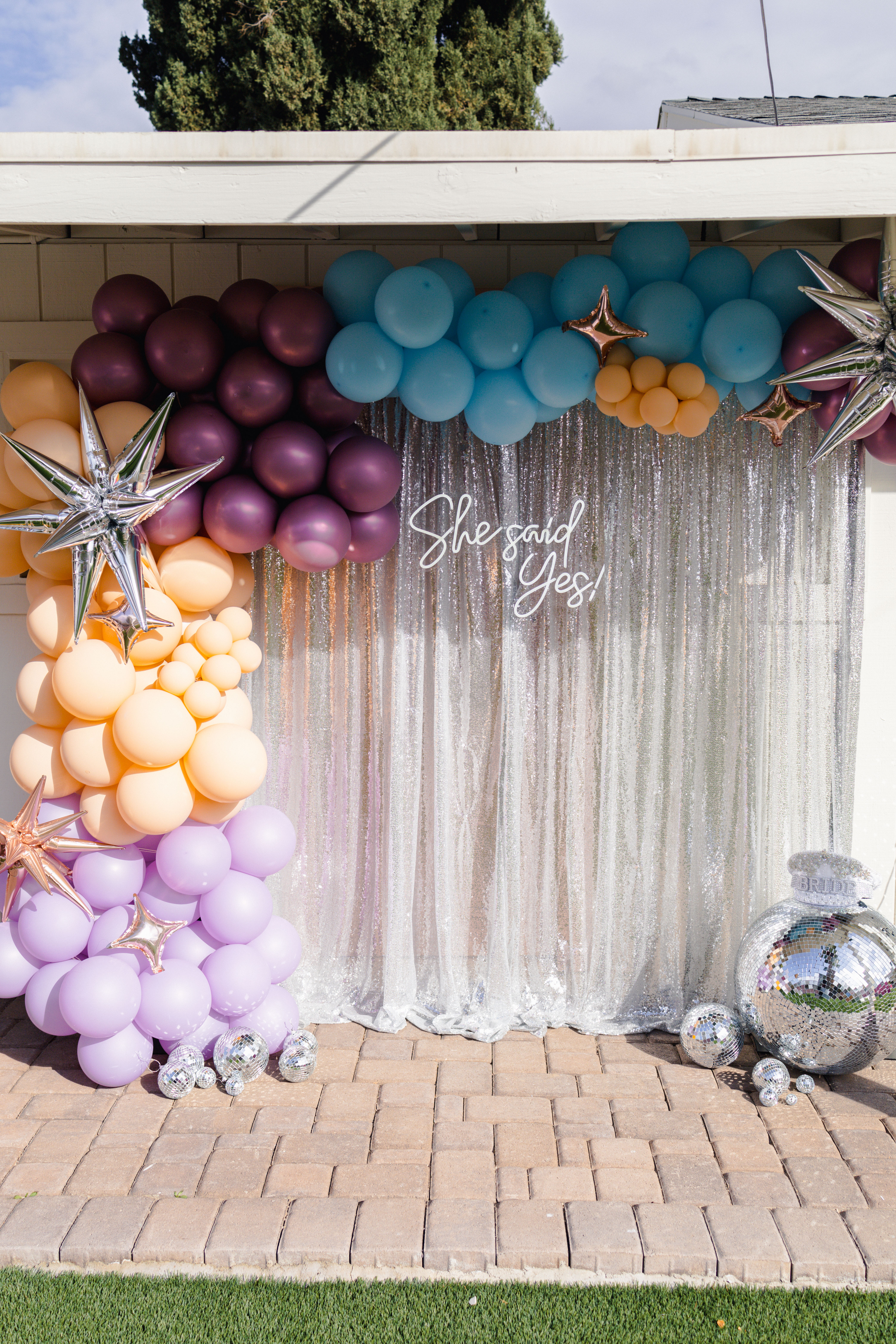 Outdoor engagement backdrop on a patio: pastel balloon arch (lavender, peach, teal, plum), metallic star balloons, disco balls and a silver sequin curtain reading She said Yes!