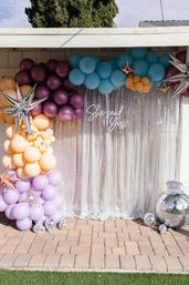 Outdoor engagement backdrop on a patio: pastel balloon arch (lavender, peach, teal, plum), metallic star balloons, disco balls and a silver sequin curtain reading She said Yes!