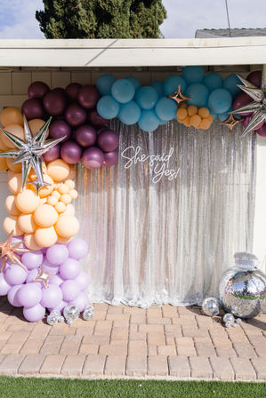 Outdoor engagement backdrop on a patio: pastel balloon arch (lavender, peach, teal, plum), metallic star balloons, disco balls and a silver sequin curtain reading She said Yes!