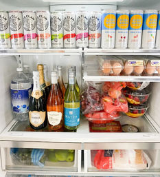 Open kitchen refrigerator stocked with canned seltzers and vodka sodas, bottles of sparkling wine and wine, club soda, eggs, containers of cut fruit and produce drawers with limes — party-ready.