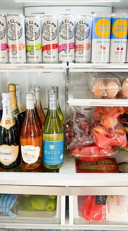 Open kitchen refrigerator stocked with canned seltzers and vodka sodas, bottles of sparkling wine and wine, club soda, eggs, containers of cut fruit and produce drawers with limes — party-ready.