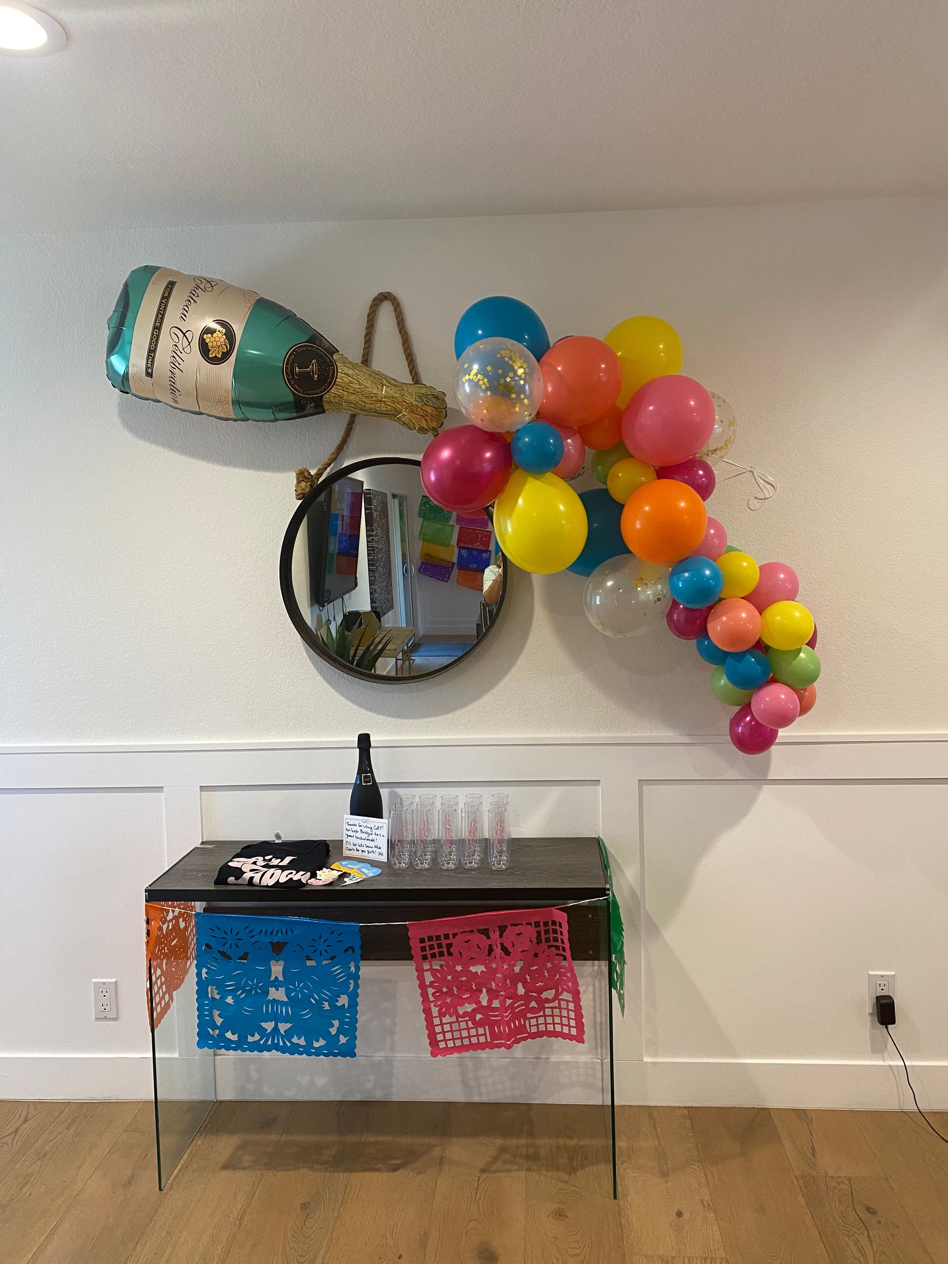 Indoor party setup in a modern entryway: oversized champagne balloon pouring a colorful balloon garland over a round mirror and console table with plastic flutes and papel picado banners.