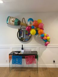 Indoor party setup in a modern entryway: oversized champagne balloon pouring a colorful balloon garland over a round mirror and console table with plastic flutes and papel picado banners.