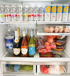 Open refrigerator stocked for a gathering: top shelf of canned hard seltzers and vodka sodas, middle shelf with sparkling and still wine bottles, club soda, carton of eggs, bags of grapes and apples, stacked fruit bowls, and lower drawers with limes and shredded cheese.