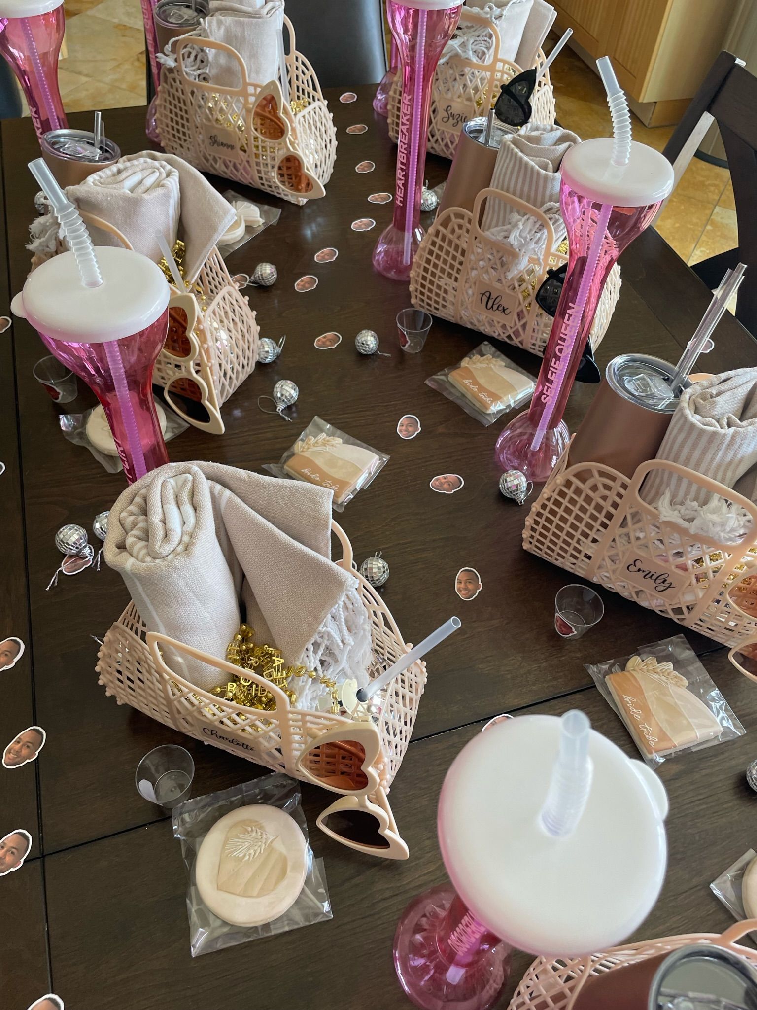 Spa-themed party table with pastel favor baskets holding rolled towels, sunglasses, treats and mini disco balls, surrounded by tall pink yard cups on a dark wood dining table.