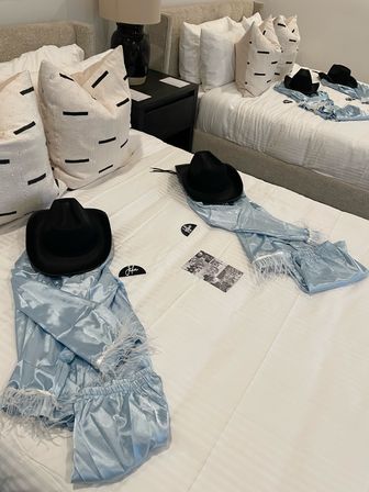 Two twin beds in a neutral guest room with white linens and patterned throw pillows, each bed styled with a black cowboy hat and light blue satin robe with feather trim on the duvet.
