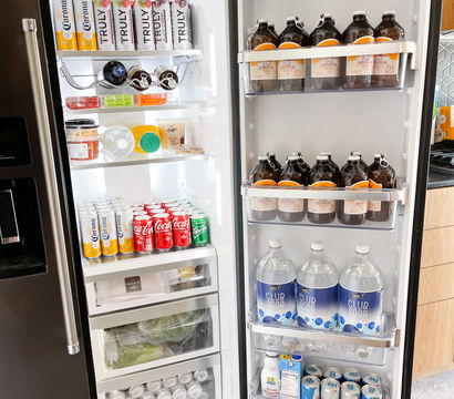 Open stainless-steel refrigerator in a modern kitchen, neatly stocked with canned seltzers and sodas (Corona, Truly, Coca-Cola, Sprite), brown glass growlers, large club soda bottles, produce drawers and bottled water — organized and party-ready.